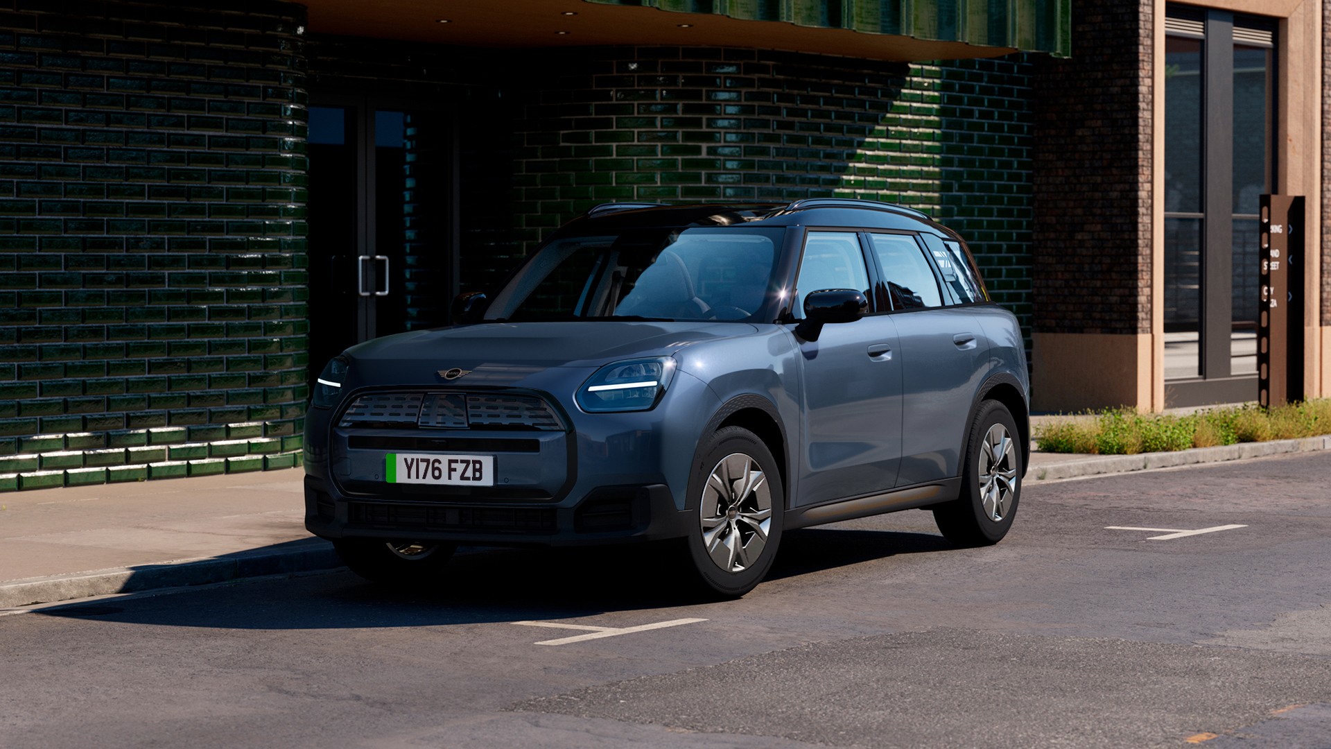 MINI Countryman electric SUV parked on a city street in front of a dark green tiled building, featuring black roof and modern headlights.